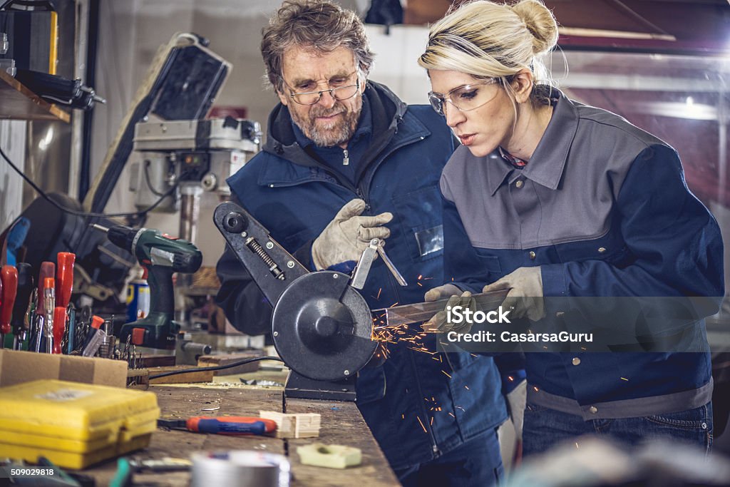 Young woman gridning metal in mechanical workshop, senior man supervising woman and teaching, employee training